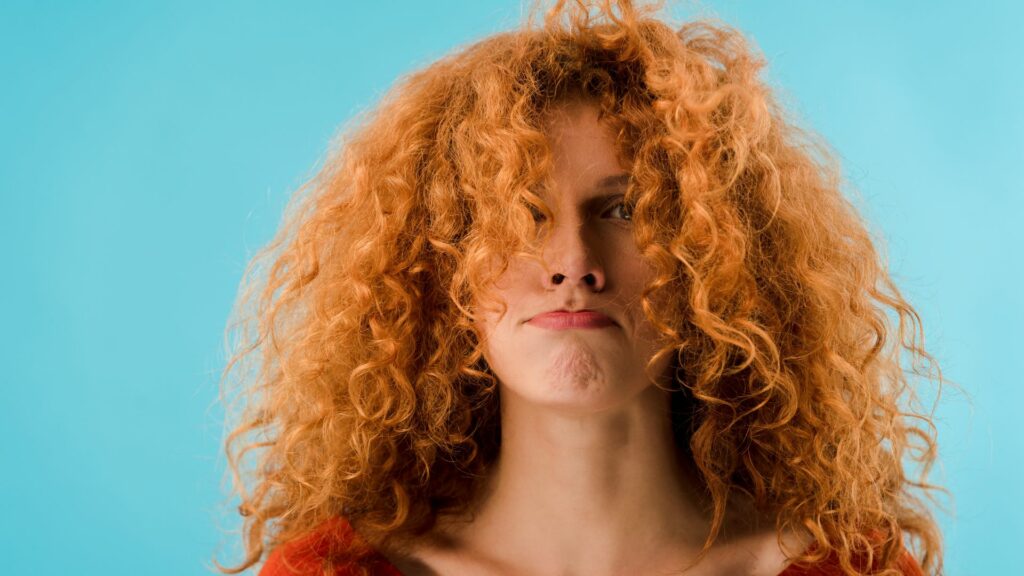 Individual with curly red hair partially obscuring their face stands before a blue background, wearing a slightly exasperated expression—ideal for an anti-frizz hair guide.