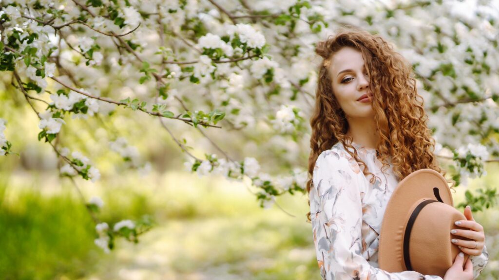 A woman with curly, lighter hair holds a hat and stands outside before tree branches adorned with blossoming white flowers, savouring the charm of brighter days.