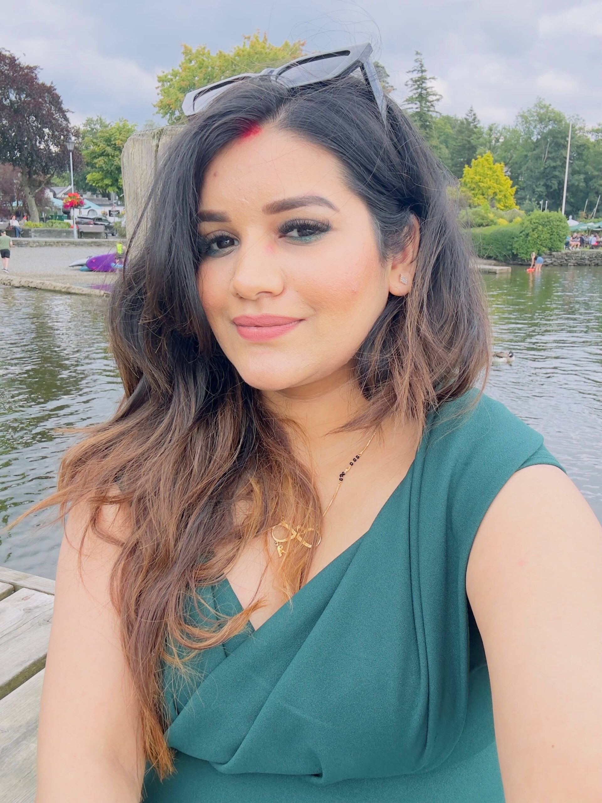 Monika, a Senior Therapist with long dark hair, wearing a green dress and sunglasses, takes a selfie by a lakeside where trees and people can be seen in the background.