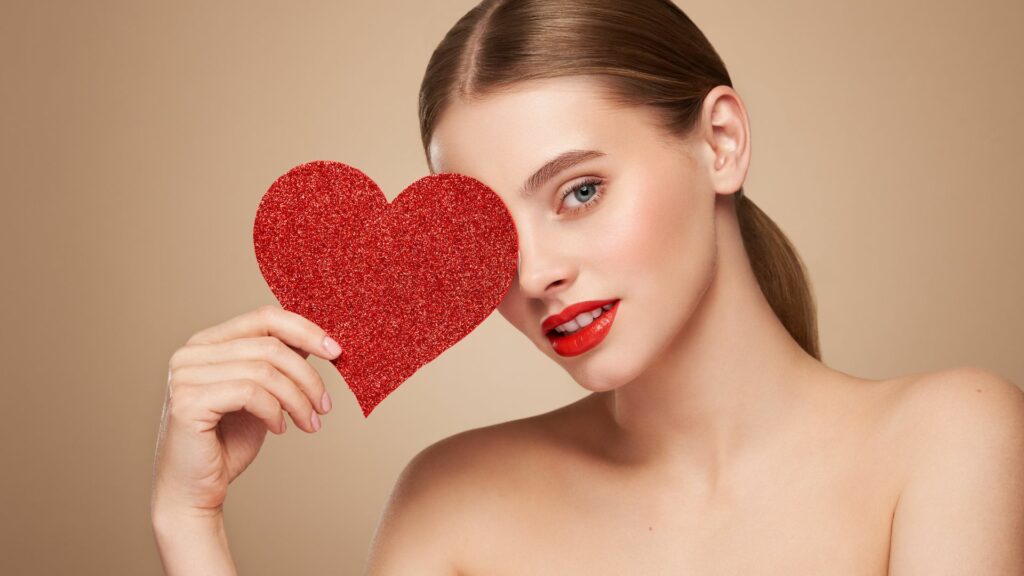 A woman wearing red lipstick holds a sparkling red heart over one eye, posing against a beige backdrop—prepared to fall in love this Valentine’s Day.