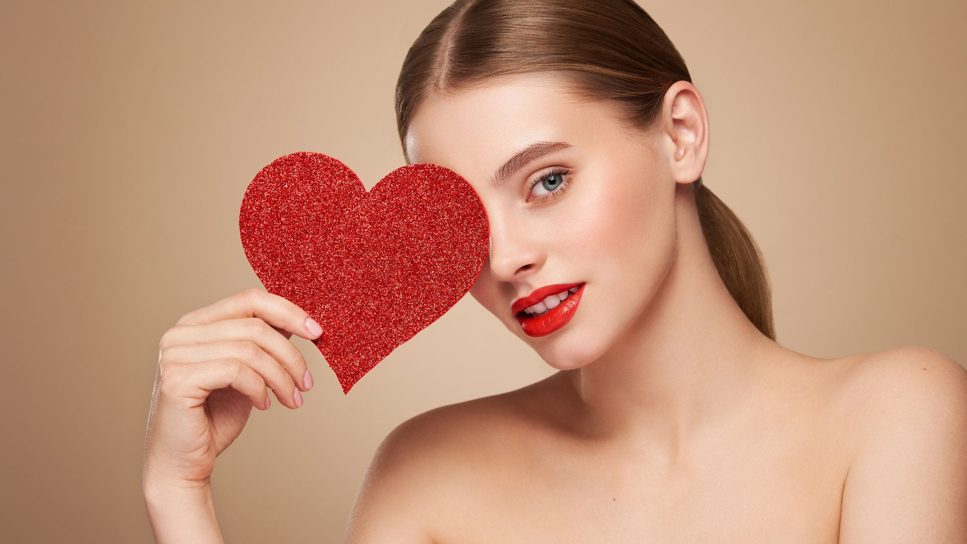 A woman wearing red lipstick holds a sparkling red heart over one eye, posing against a beige backdrop—prepared to fall in love this Valentine’s Day.