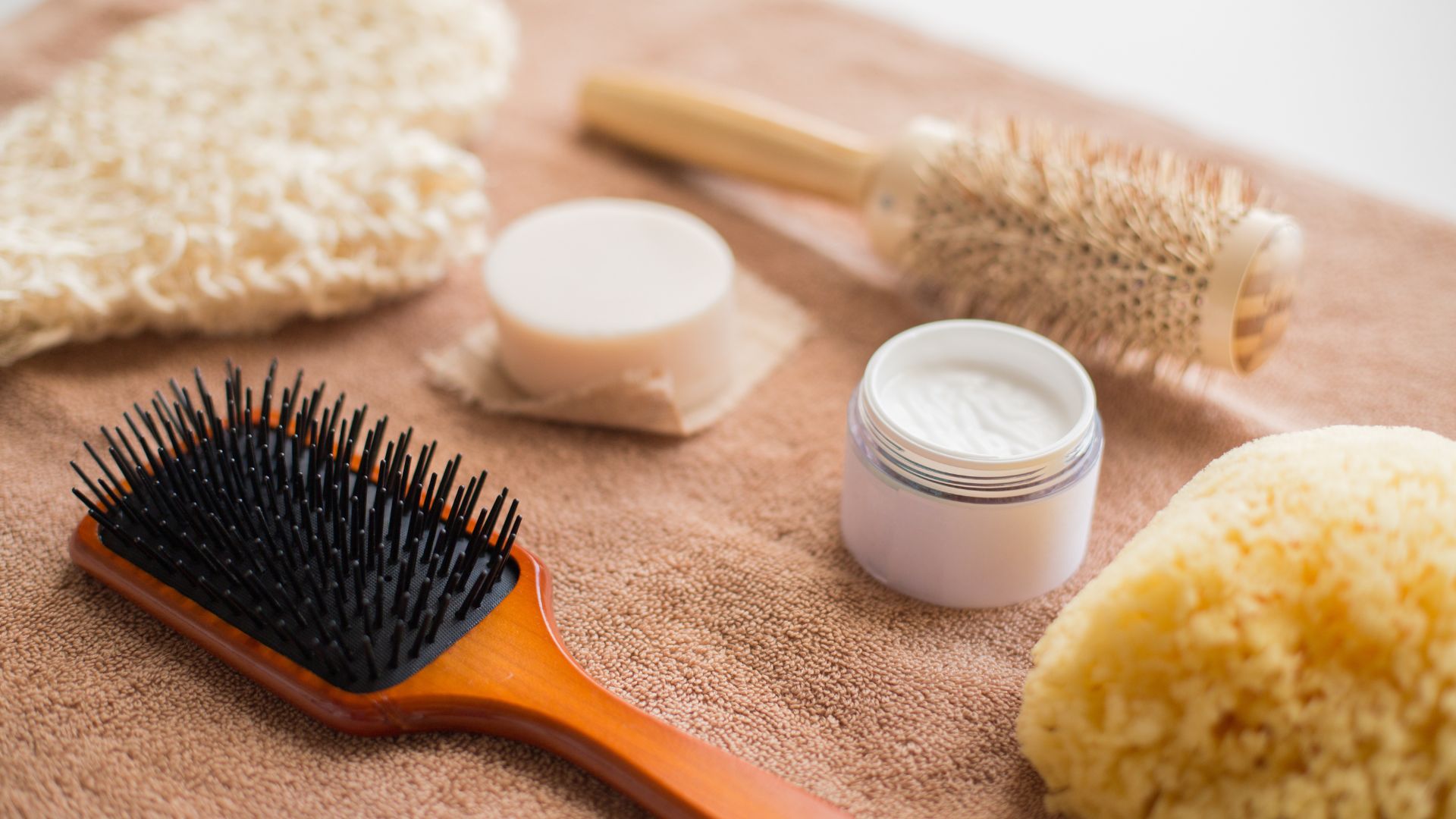 A wooden hairbrush, a round brush, a jar of cream, and other essentials for humidity-resistant hair are laid out on a brown towel, ready to help you achieve a smooth, frizz-free look.