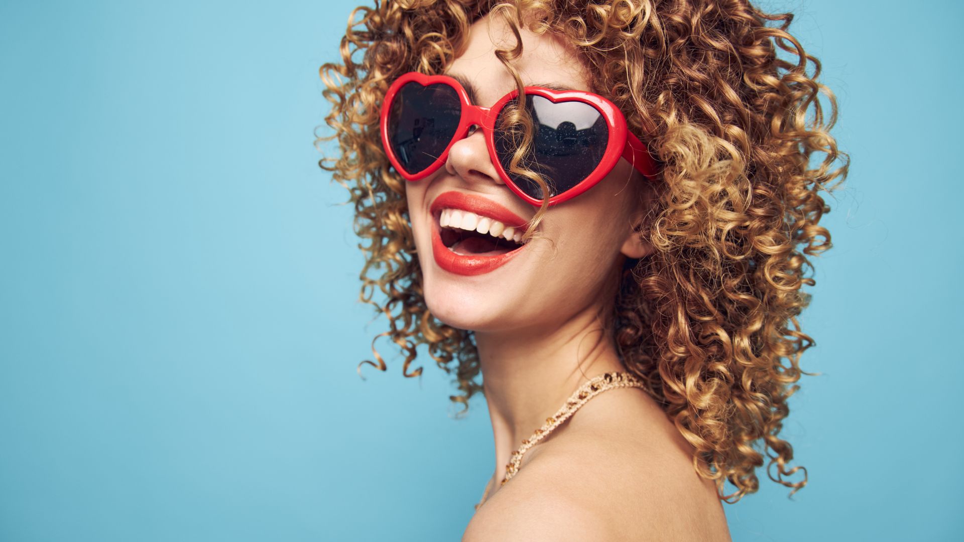 Adore your style as this woman with curly hair, red heart-shaped sunglasses, and a gold necklace beams against a simple blue background.