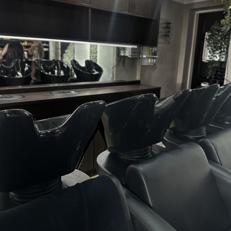 A row of black salon shampoo chairs faces a counter and large mirror; two people can be seen reflected in the background.