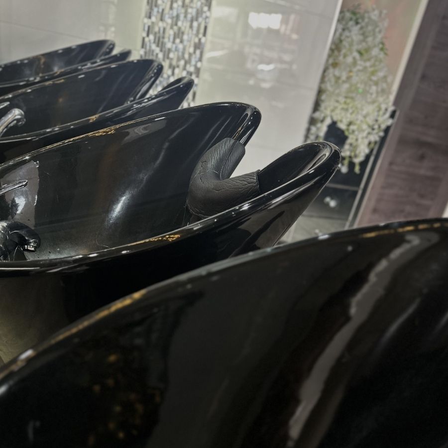 A row of black salon wash basins with neck supports, set before a tiled wall with a decorative plant in the background.