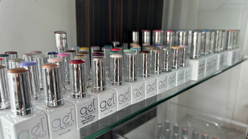 A row of gel nail varnish bottles with silver lids is arranged on a glass shelf, each bottle marked 'gel bottle' and displaying various colours on the top.