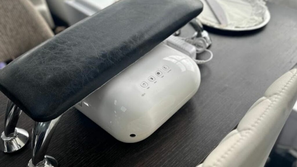 A black padded armrest and a white nail dryer with control buttons are set on a dark wooden table beside a white chair.