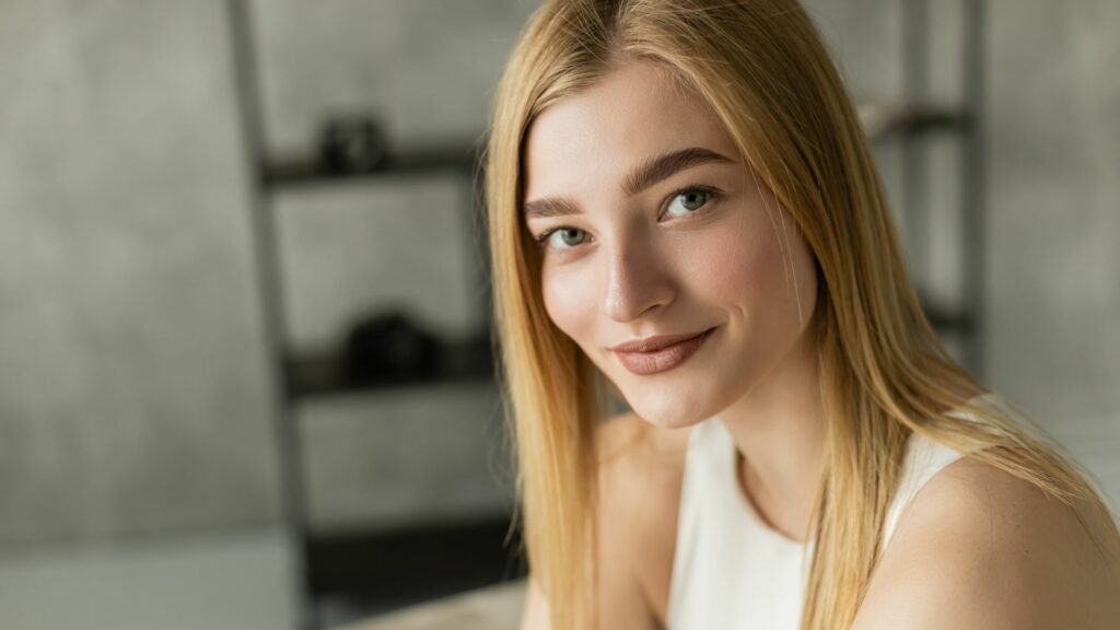 A young woman with straight blonde hair, a low-maintenance hairstyle, and fair skin wears a sleeveless white top and smiles at the camera, seated indoors with a blurred shelf in the background.
