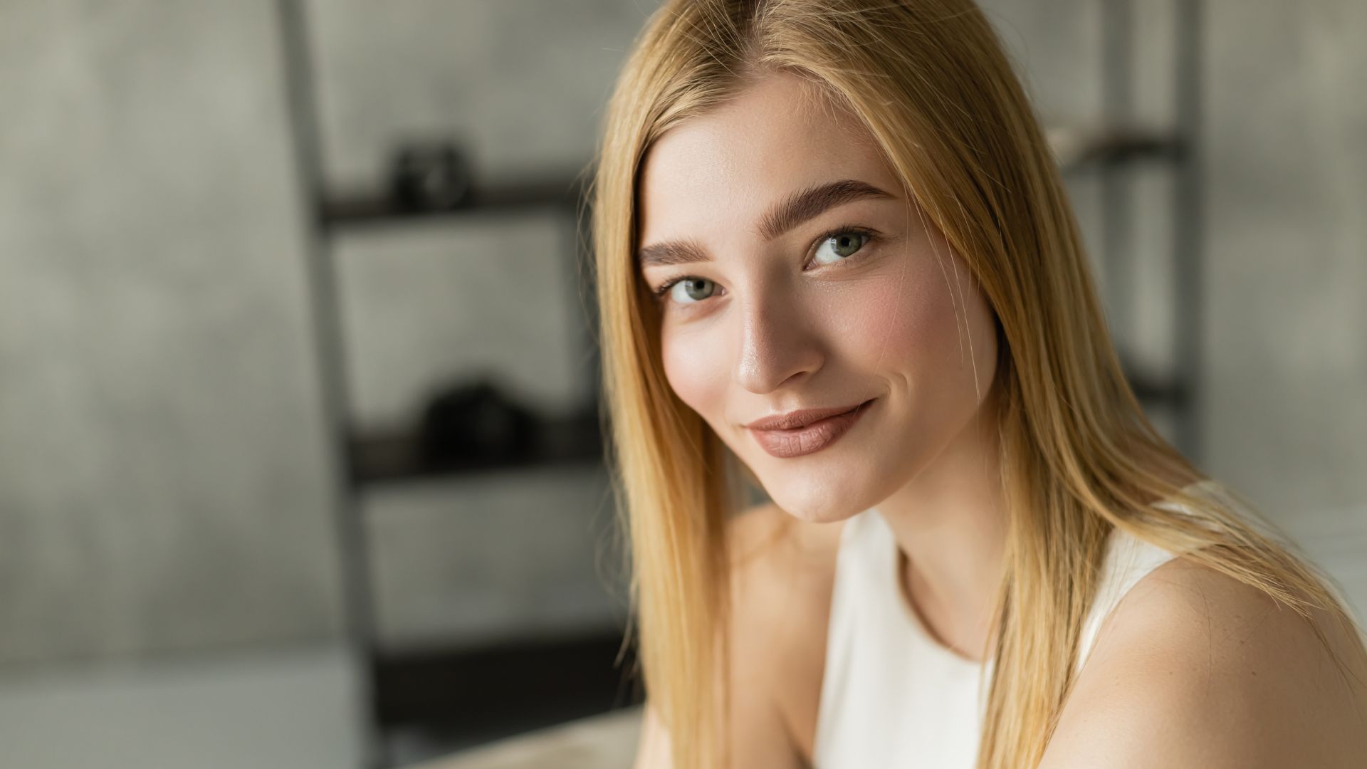 A young woman with straight blonde hair, a low-maintenance hairstyle, and fair skin wears a sleeveless white top and smiles at the camera, seated indoors with a blurred shelf in the background.