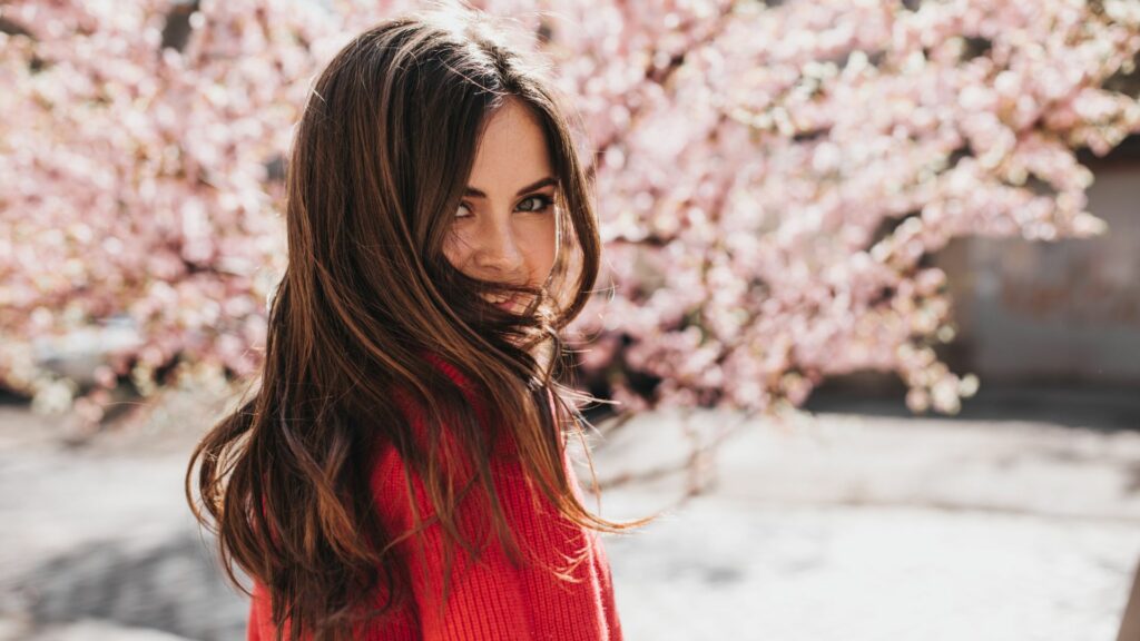 A woman with long brown hair, dressed in a red jumper, stands outside before blooming pink cherry blossom trees, welcoming the new season as she glances back over her shoulder.