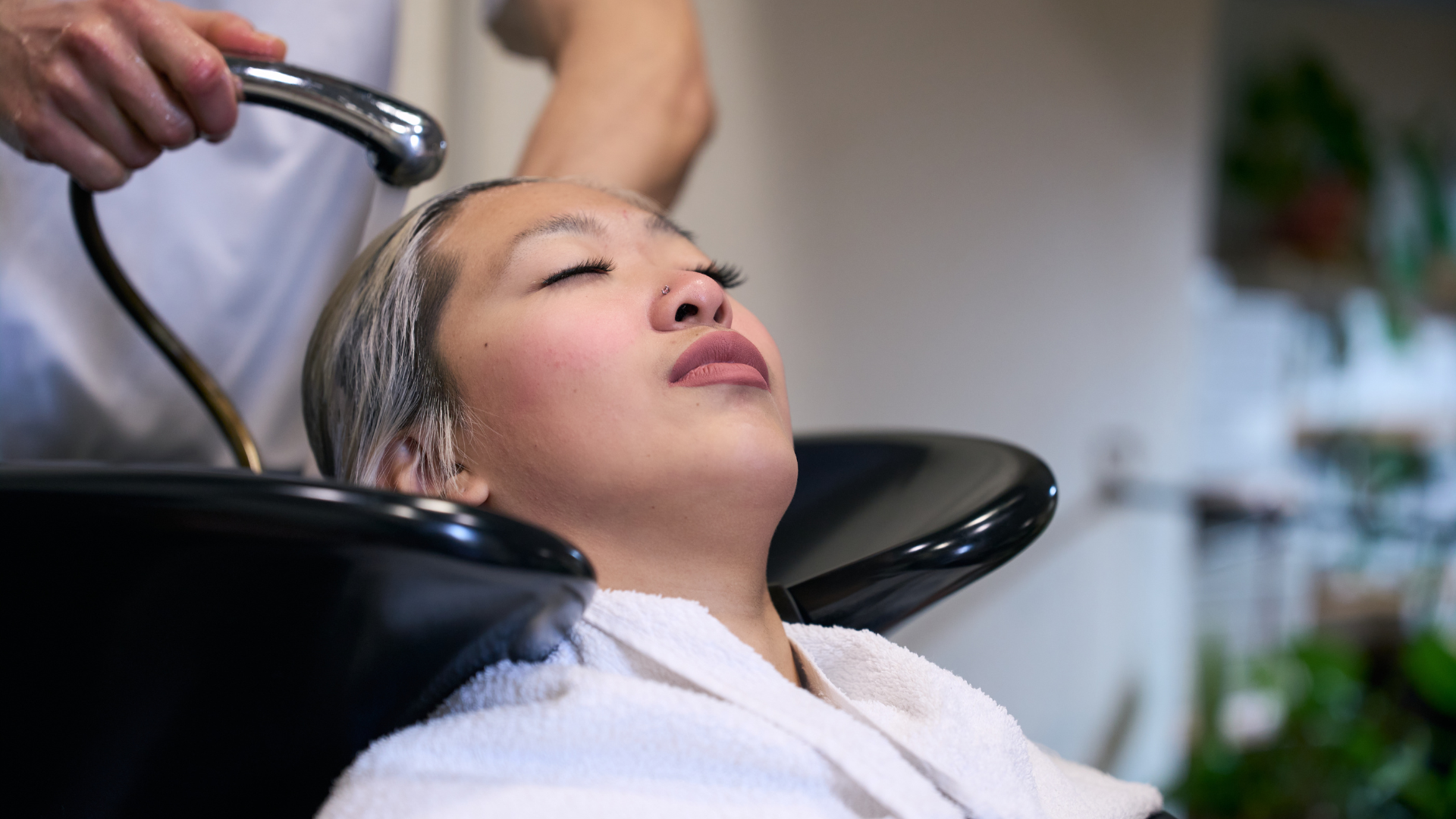 A person with eyes closed reclines in a salon chair as their stylist gives a soothing scalp massage and hair treatment, washing their hair under a basin for healthy tresses.
