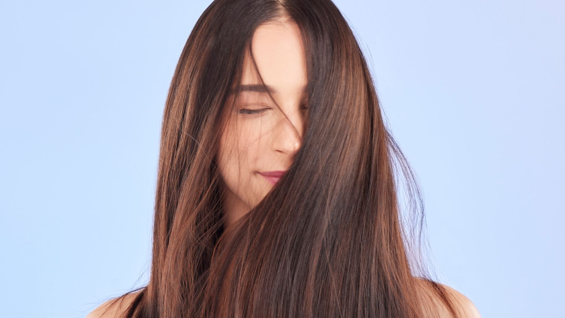 A woman with long, straight brown hair stands before a light blue background, her eyes closed and hair partly covering her face—highlighting a classic hairstyle that needs minimal maintenance.