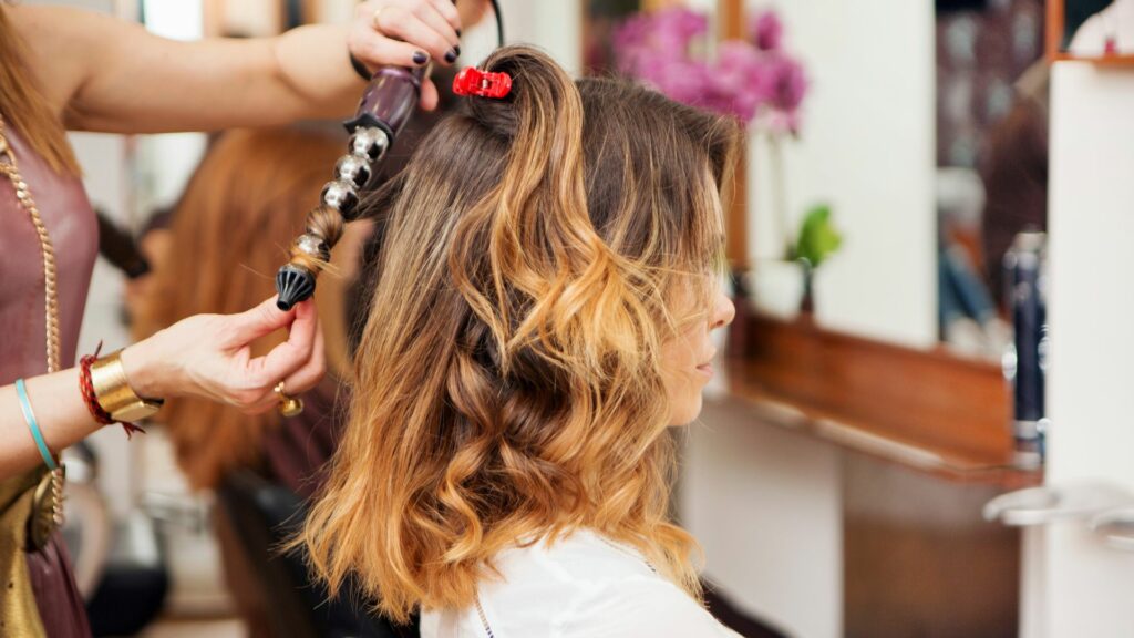 A hairdresser styles a woman's shoulder-length hair with a curling tong in a salon. The woman sits sideways, enjoying soothing salon treatments as part of her Bank Holiday beauty routine.