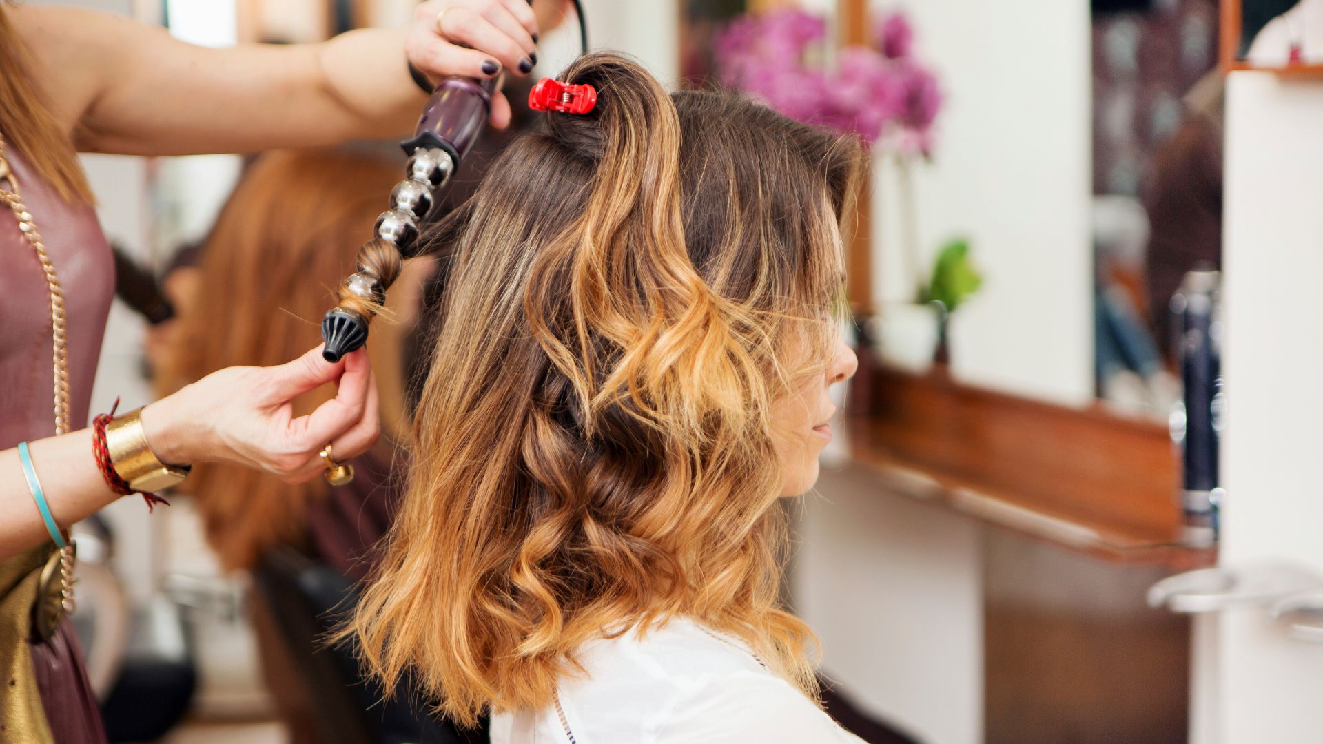 A hairdresser styles a woman's shoulder-length hair with a curling tong in a salon. The woman sits sideways, enjoying soothing salon treatments as part of her Bank Holiday beauty routine.