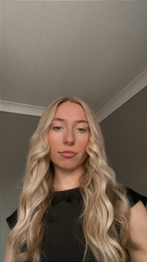 An individual with long, wavy blonde hair dressed in a black top stands indoors against a backdrop of plain walls and a white ceiling.