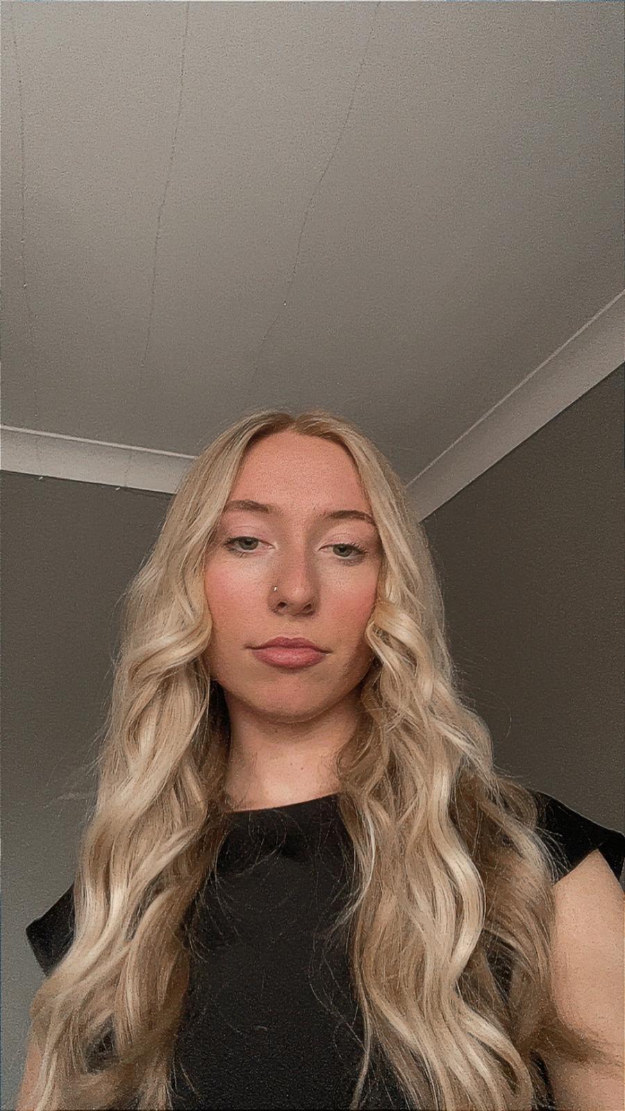 An individual with long, wavy blonde hair dressed in a black top stands indoors against a backdrop of plain walls and a white ceiling.