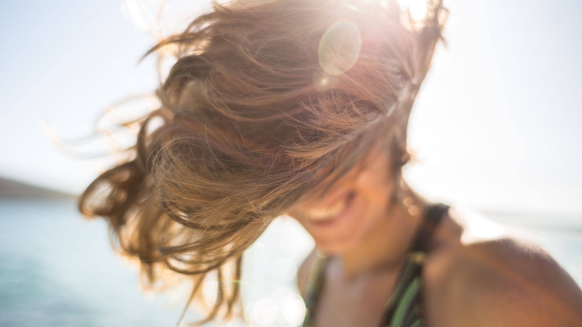 A person with sunlit brown hair falling over their face smiles outdoors by the water on a bright day.