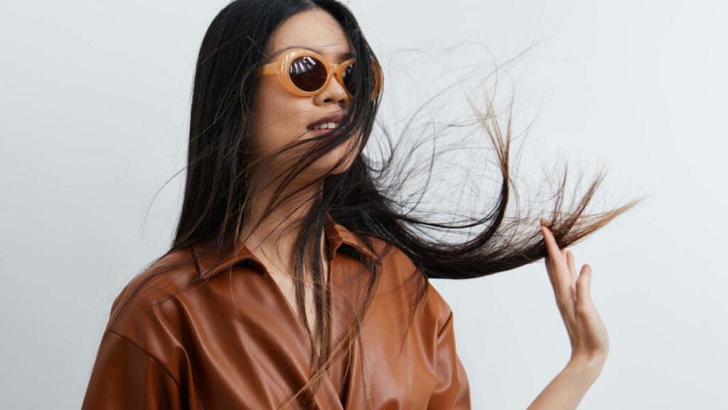 A woman in brown sunglasses and a brown leather shirt flicks her long dark hair with stylish flair; her summery look is striking against a simple, pale background.