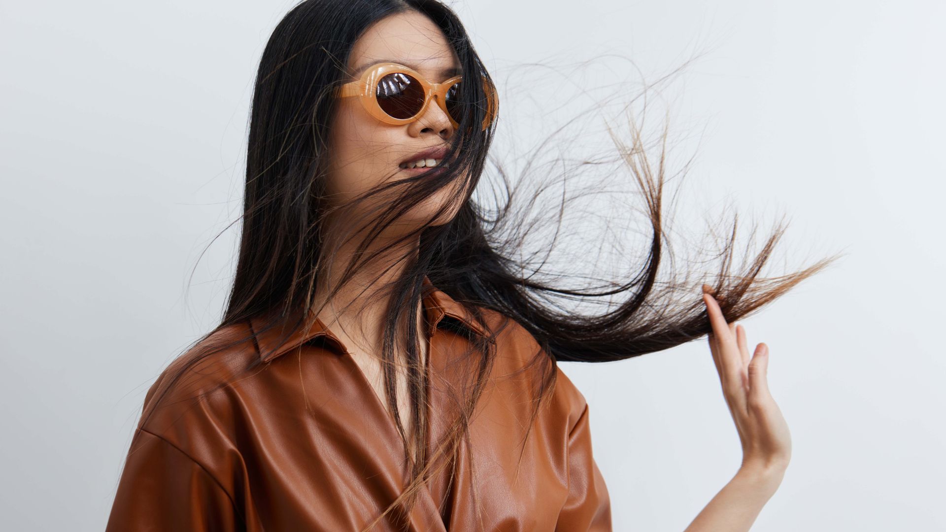 A woman in brown sunglasses and a brown leather shirt flicks her long dark hair with stylish flair; her summery look is striking against a simple, pale background.