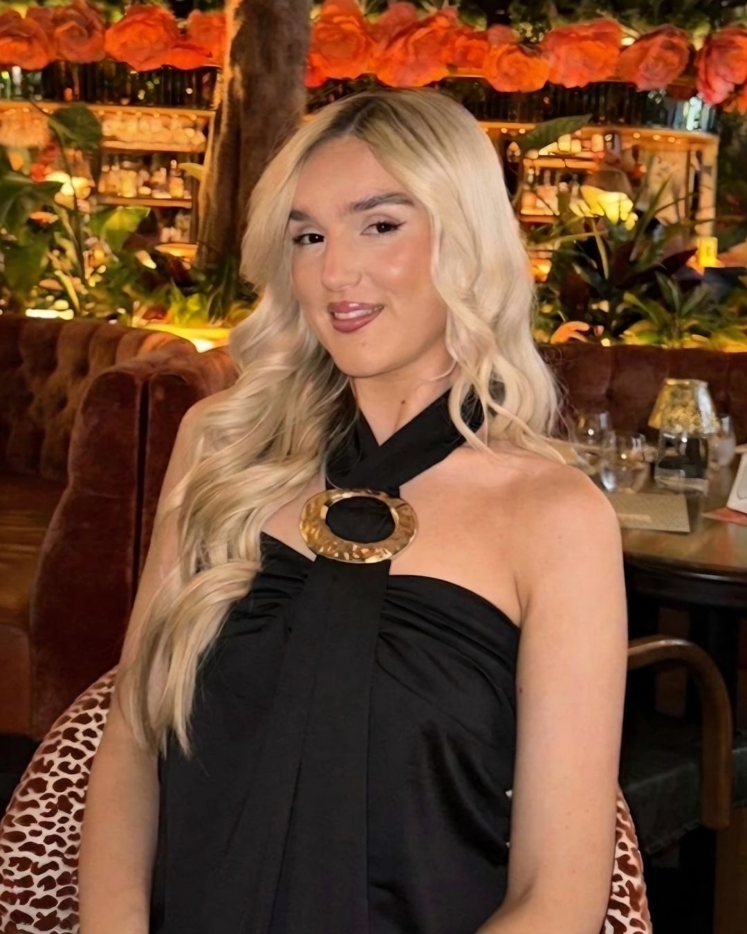 A person with long blonde hair in a black halter dress is seated at a restaurant featuring floral decorations and subdued lighting.