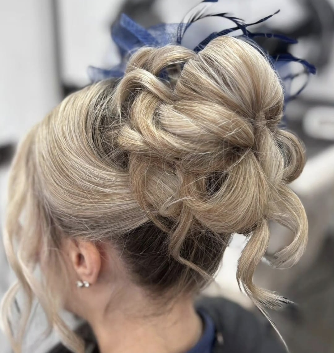 A woman with blonde hair arranged in an elaborate updo featuring loose curls and adorned with a blue feathered accessory, highlighting the refined look of a macadamia hair spa treatment in Widnes.