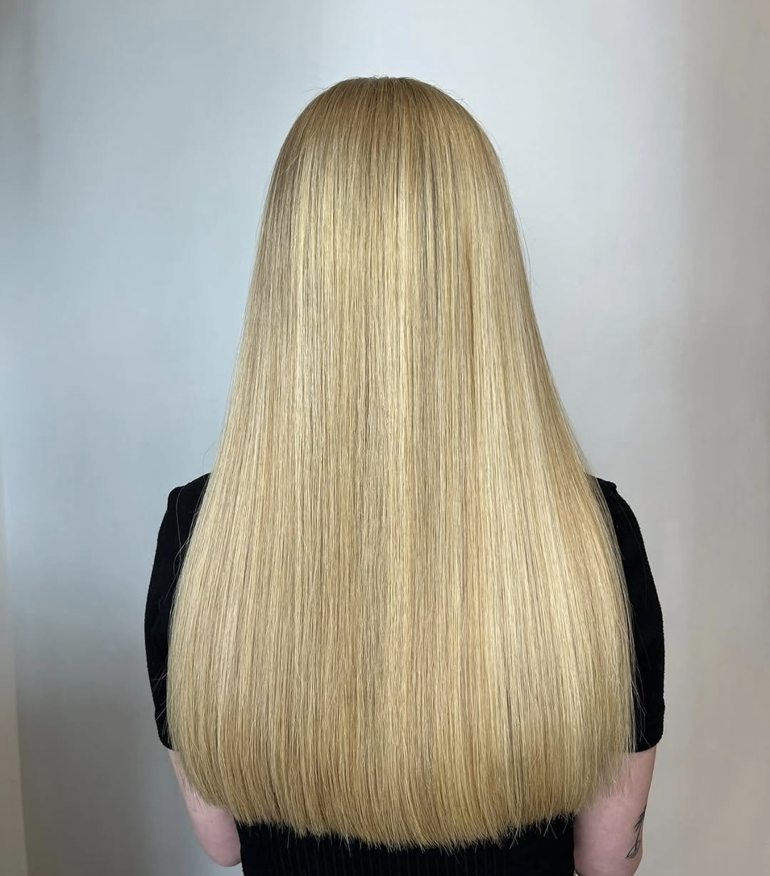Individual with sleek, long blonde hair—expertly styled using hair smoothing Widnes—dressed in a black shirt, seen from behind against a simple light-coloured background.