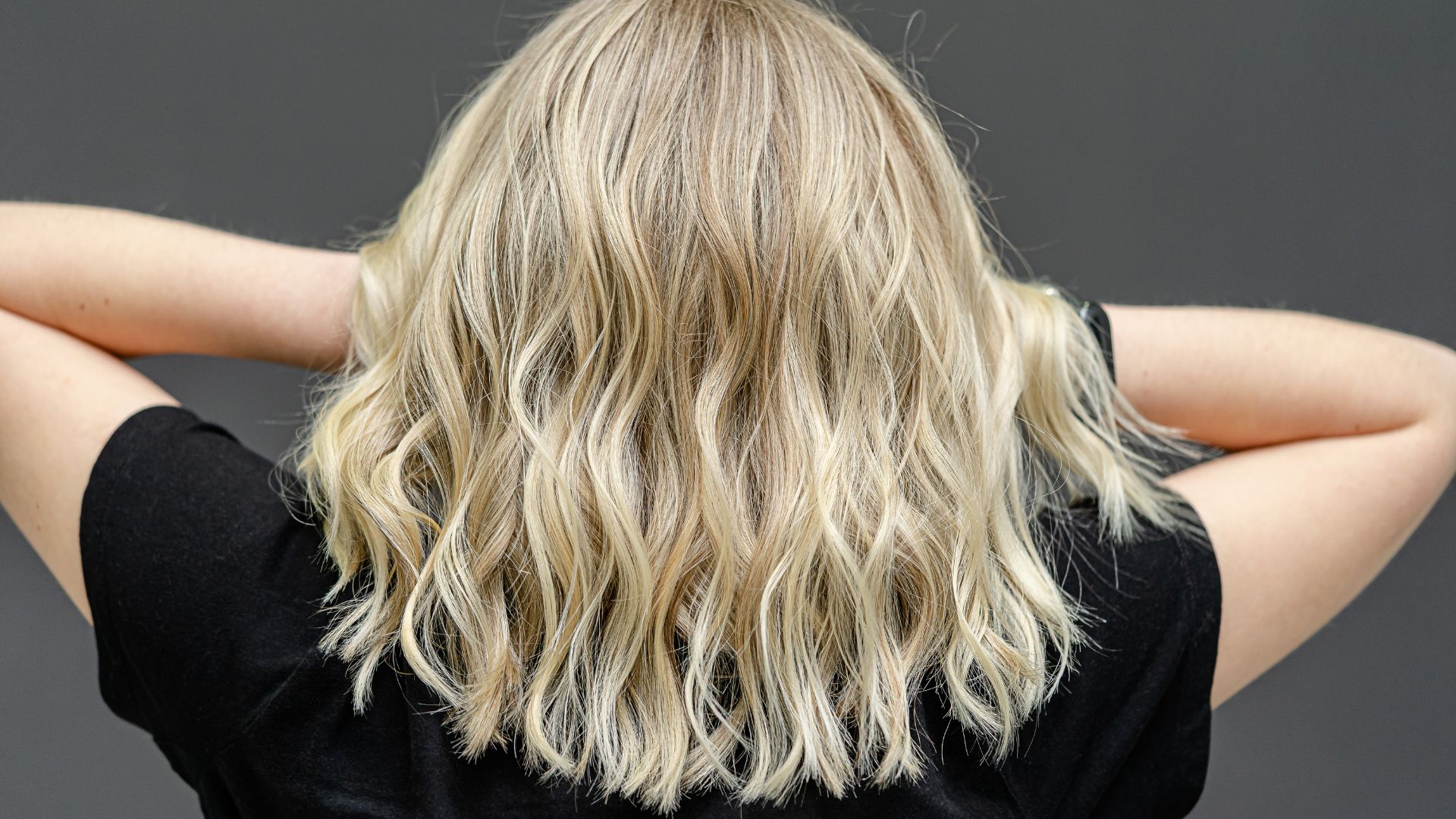 Individual with wavy, shoulder-length blonde hair seen from behind, seated on a chair in a black shirt, hands placed behind their head as though pausing for a moment of relaxation.