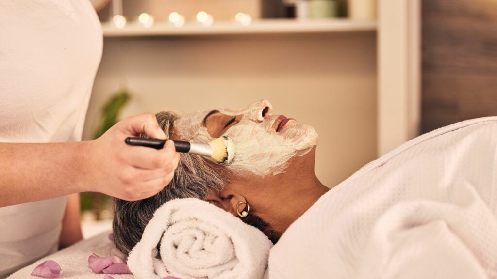 A person lies on a spa bed with a towel beneath their head whilst a spa therapist applies a bespoke facial mask using a brush.