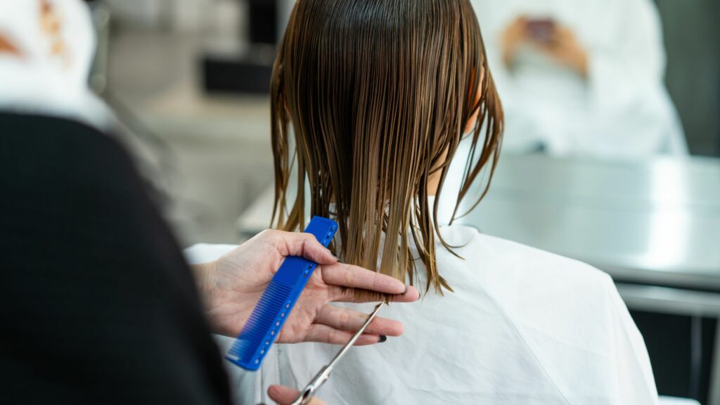 A person with wet hair is having their hair cut; a hand trims the hair in a straight line at the back using scissors and a blue comb, much like how a bespoke facial adapts treatments to personal requirements.