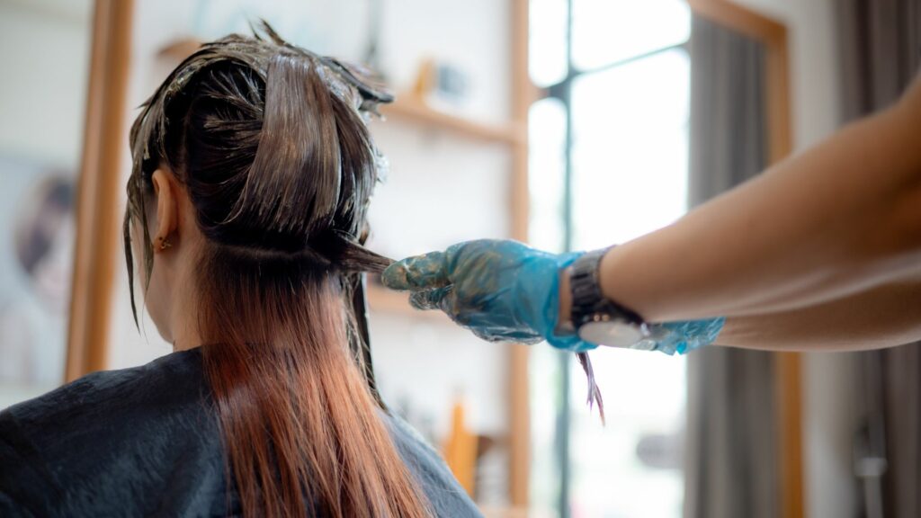 A person with long hair sits as another, wearing gloves, applies dye to the back of their head in a salon, ensuring beautifully coloured hair.