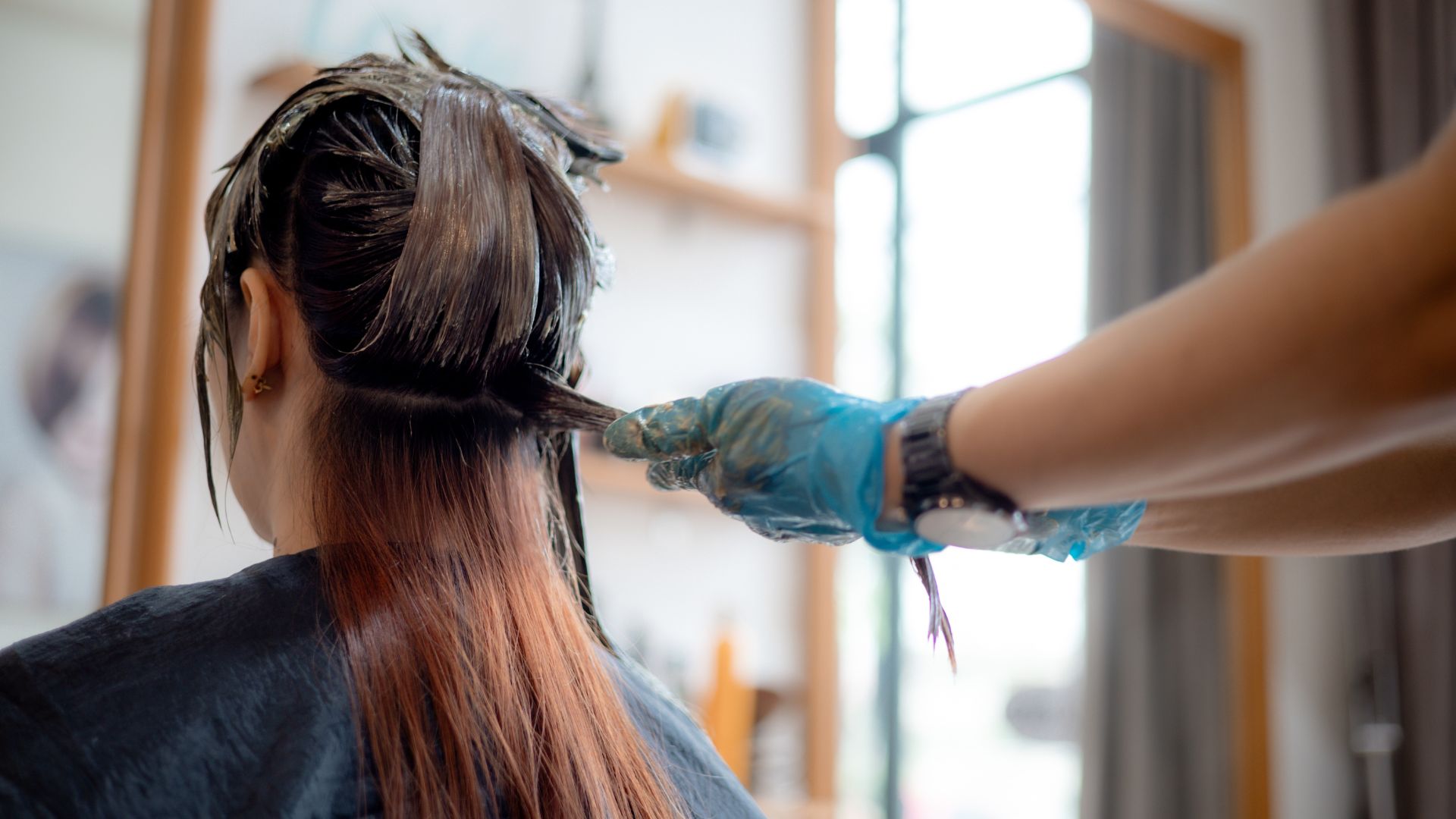 A person with long hair sits as another, wearing gloves, applies dye to the back of their head in a salon, ensuring beautifully coloured hair.