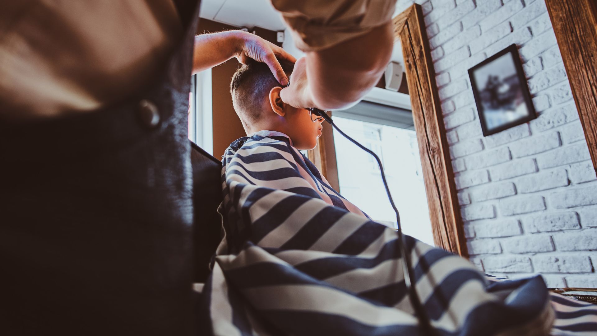 A barber from Macadamia Hair Spa Barber Services is cutting a young boy’s hair with clippers in the salon, while the boy wears a striped barber’s gown.