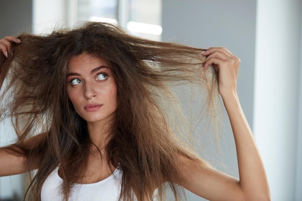 A woman with long brown hair in a white vest top lifts sections of her frizzy, tangled hair, gazing sideways and pondering the need for post-holiday hair recovery in a brightly lit room.
