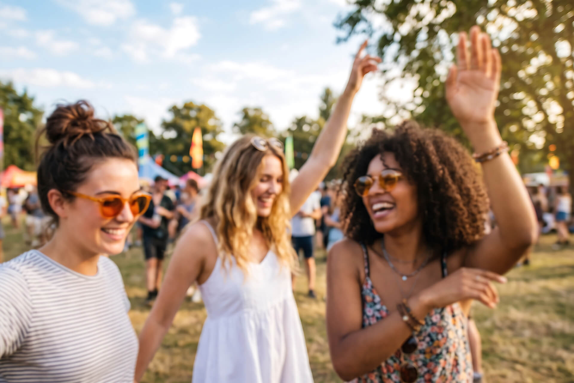 Three women enjoying themselves at a festival, smiling and dancing in the sunshine, displaying bright festival hairstyles, with a crowd and trees visible behind them.