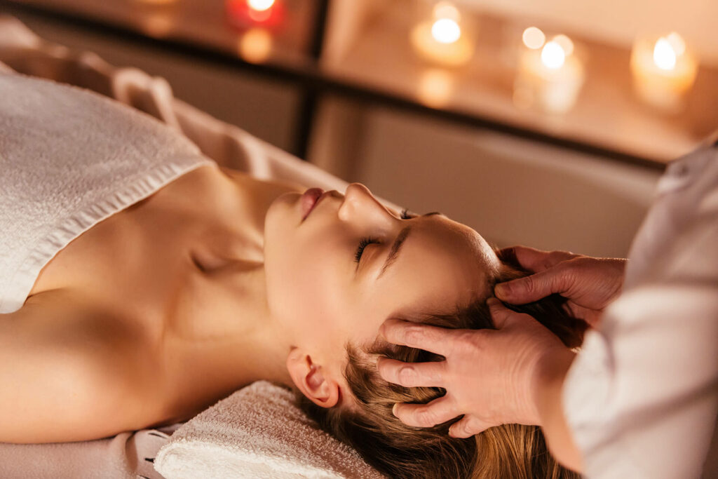 Someone lies on a massage table with eyes closed, relishing a calming scalp treatment as another person massages their head. Gently glowing candles in the background create a peaceful and tranquil ambience.