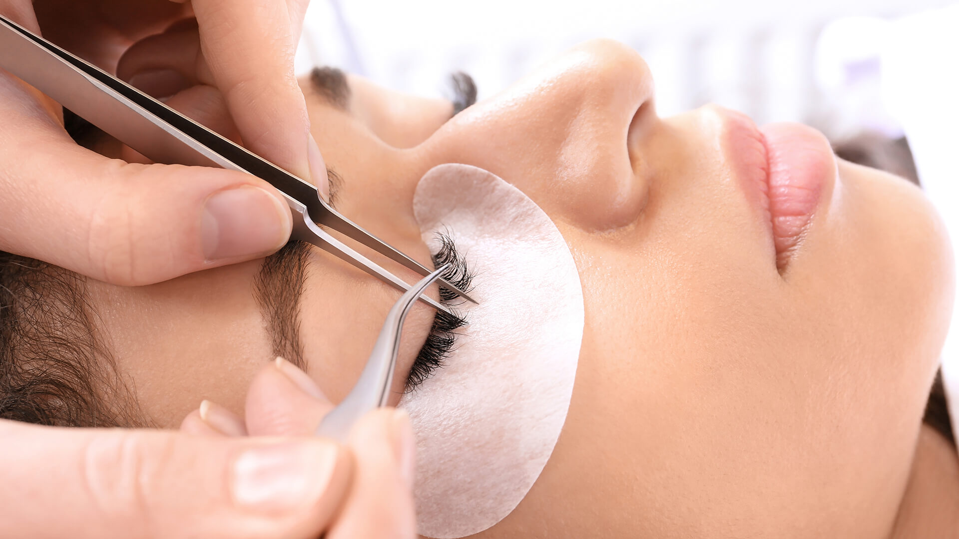 An individual reclines with closed eyes while a technician skilfully applies eyelash extensions using tweezers. A white pad beneath the eye offers protection, highlighting the newest lash trend.