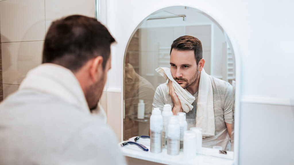 A man stands before a bathroom mirror, drying his face with a towel. Various men’s grooming products are tidily placed on the counter, highlighting a daily self-care and style routine.