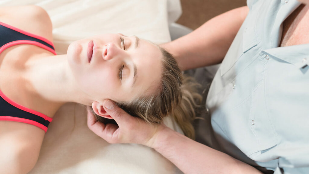 A person is lying on a table with eyes closed as another supports their head, possibly administering a Nervous System Massage—a trending wellness therapy popular for promoting deep relaxation and healing.