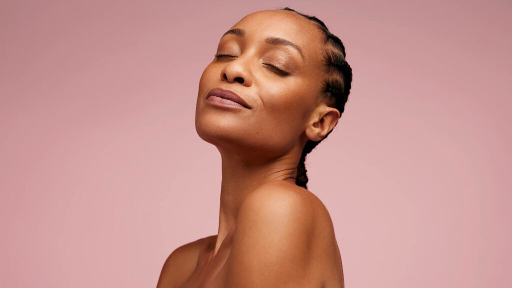 A woman with plaited hair tilts her head back and to the side, eyes closed and expression calm, showcasing a Skin-First Make-up style against a simple pink backdrop—reflecting Autumn 2026’s softer beauty trend.