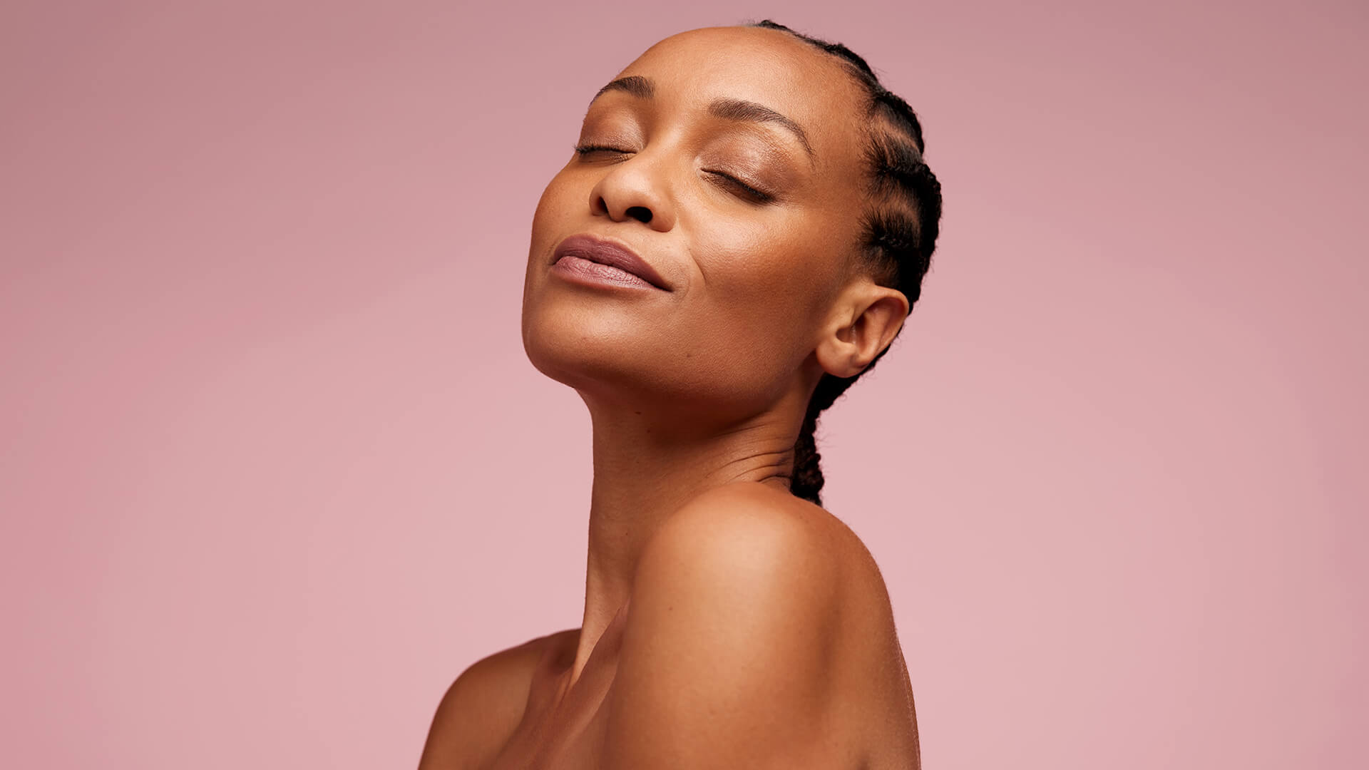 A woman with plaited hair tilts her head back and to the side, eyes closed and expression calm, showcasing a Skin-First Make-up style against a simple pink backdrop—reflecting Autumn 2026’s softer beauty trend.