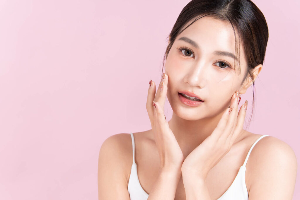 A woman with long dark hair in a white sleeveless top poses against a pale pink background, gently touching her face with both hands and displaying the radiant complexion inspired by Glass Skin Facials.