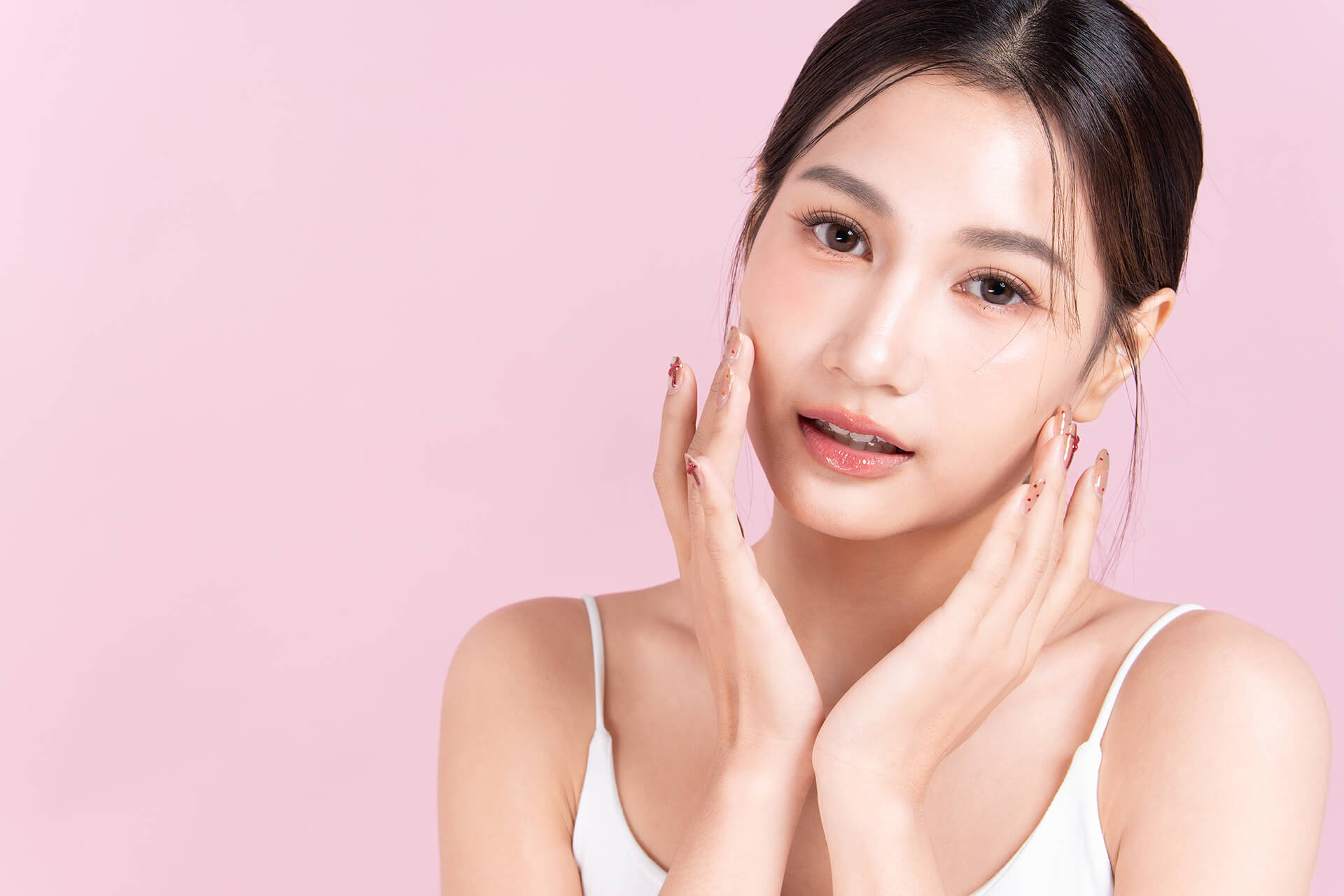 A woman with long dark hair in a white sleeveless top poses against a pale pink background, gently touching her face with both hands and displaying the radiant complexion inspired by Glass Skin Facials.