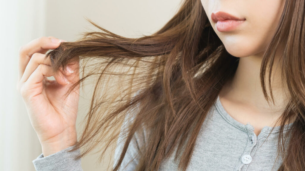 A person in a grey shirt looks at the ends of their long, brown hair with their hand, possibly thinking about hair styling tricks for skipping a wash and embracing second-day hair.