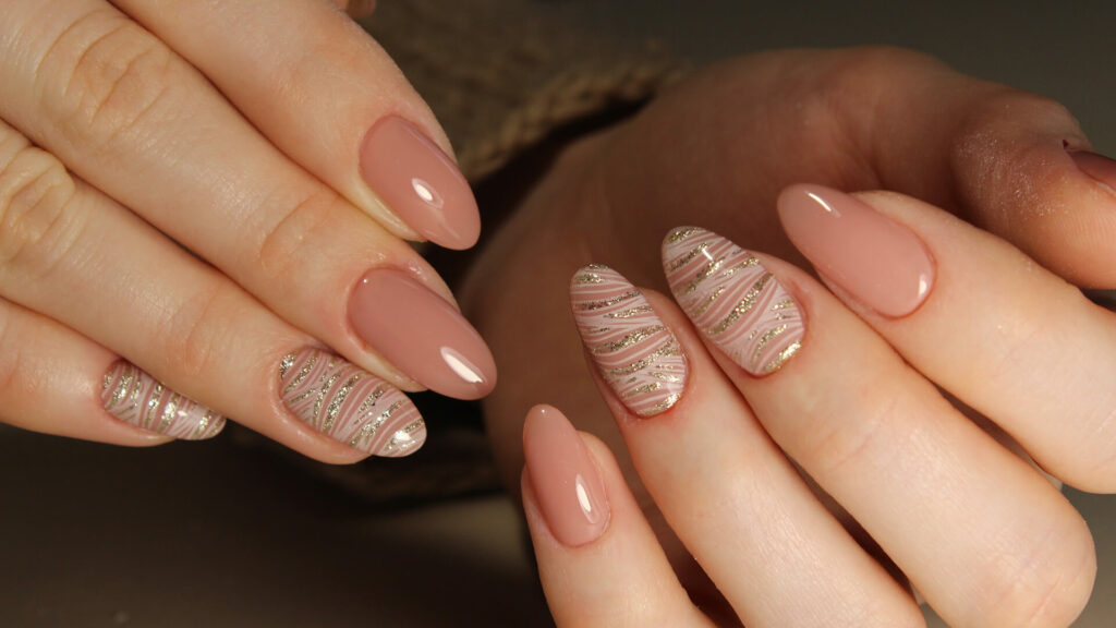 Close-up of almond-shaped nails polished in nude pink; selected nails display glittery gold and white striped designs, ideal for highlighting autumn manicure styles and the latest British nail trends.