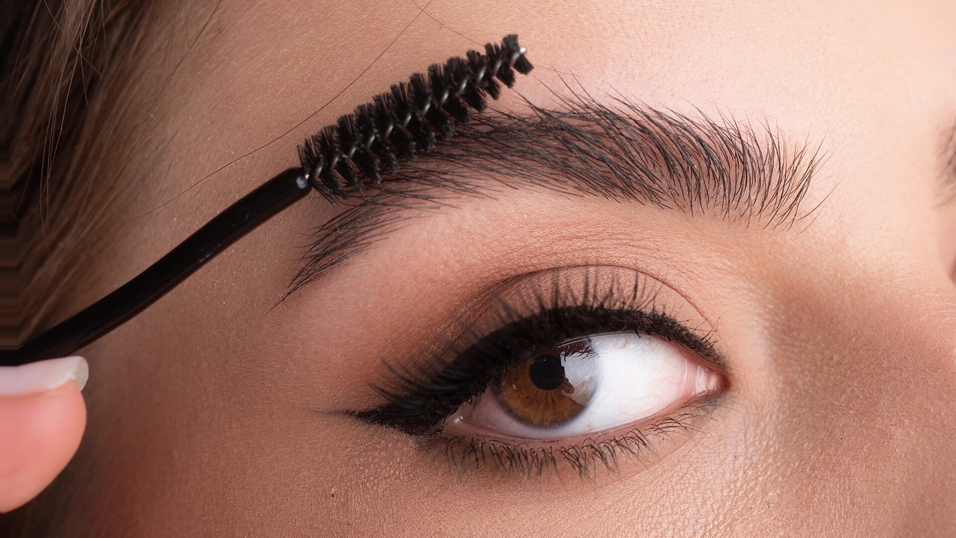 Close-up of a person grooming their eyebrow with a spoolie brush, highlighting 2026 eyebrow trends. The eye displays winged eyeliner and long lashes.