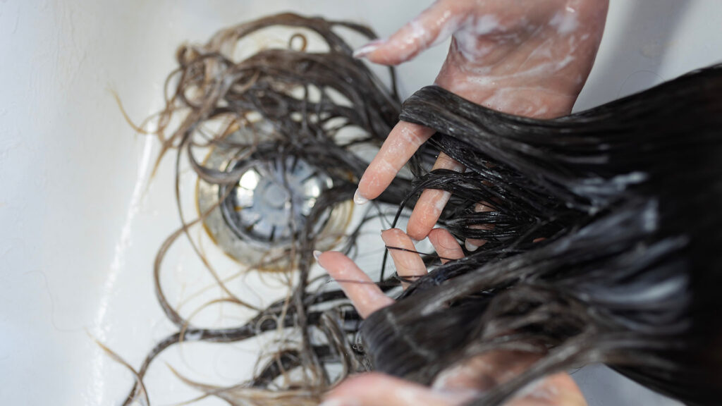 Hands washing long, wet hair in a basin, with water and soap visible on both the hair strands and hands—a typical depiction of normal hair shedding before it reaches the plughole in the shower.