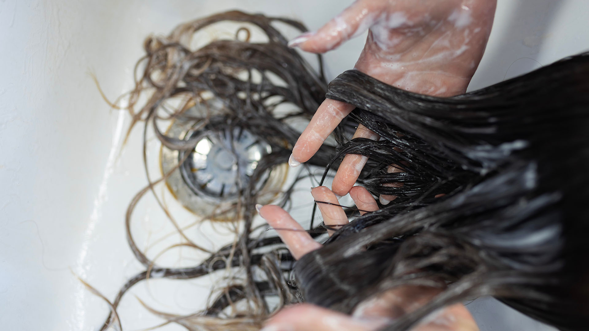 Hands washing long, wet hair in a basin, with water and soap visible on both the hair strands and hands—a typical depiction of normal hair shedding before it reaches the plughole in the shower.