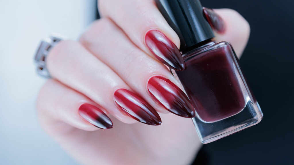 A hand with long, almond-shaped nails in a red-to-black gradient holds a matching dark red varnish bottle—ideal for Spooky-Chic Hallowe’en Nails 2026.