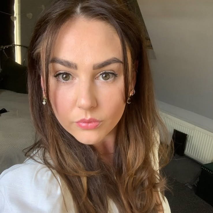 A woman with long brown hair and subtle makeup gazes straight at the camera. She is indoors, with a bed, cushions, and a radiator visible in the background.