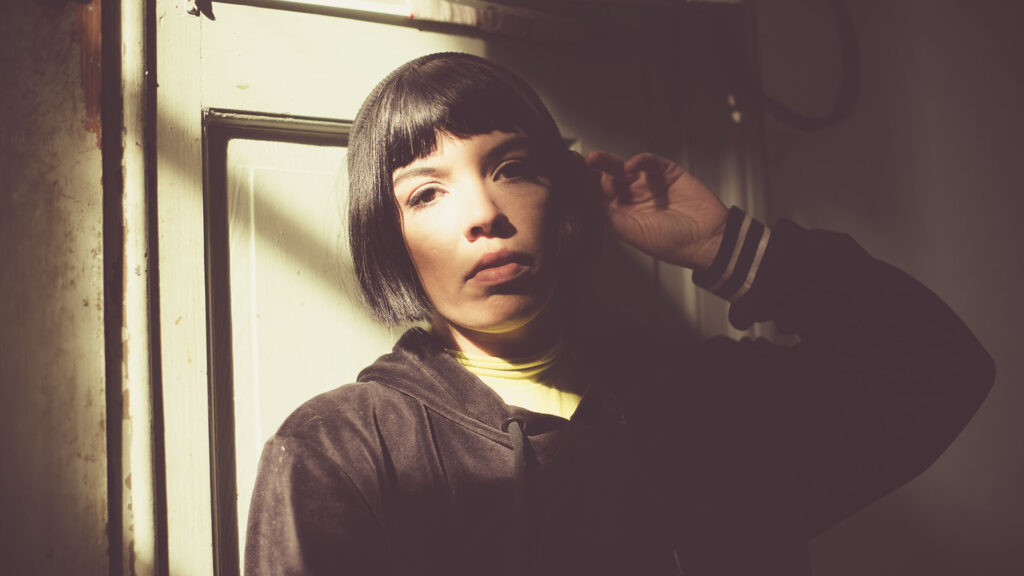 A person with a dark bob haircut, a key look for autumn hair trends, wears a black hoodie over a yellow top whilst positioned against a light-coloured door.